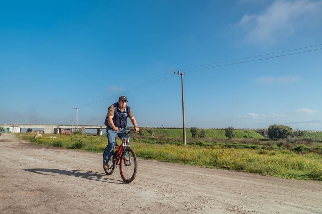 A man riding a bike on a sunny rural dirt road with a bridge and fields in the background.