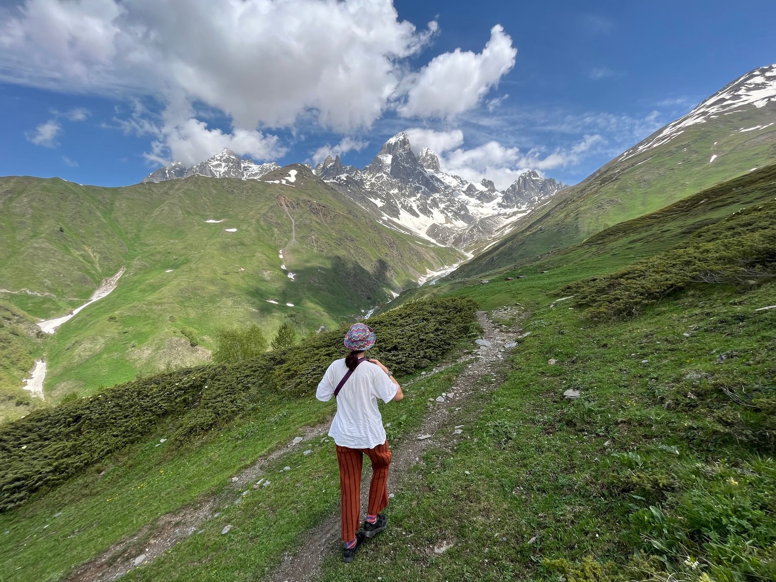 A woman walks along a trail in the breathtaking mountains of Georgia under a partly cloudy sky.